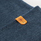 Nahaufnahme einer dunkelblauen Beanie-Mütze mit feiner Strickstruktur. Im Fokus: ein brauner Kunstleder-Patch mit Kompass-Logo und goldener Niete – nachhaltig veredelt in Deutschland, erhältlich bei nordkit.de.