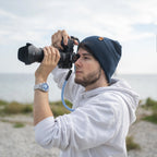 Junger Mann mit dunkelblauer Beanie-Mütze und weißem Hoodie fotografiert mit einer Spiegelreflexkamera am Meer. Die Mütze mit Kompass-Patch aus Kunstleder wurde nachhaltig veredelt in Deutschland – erhältlich bei nordkit.de.