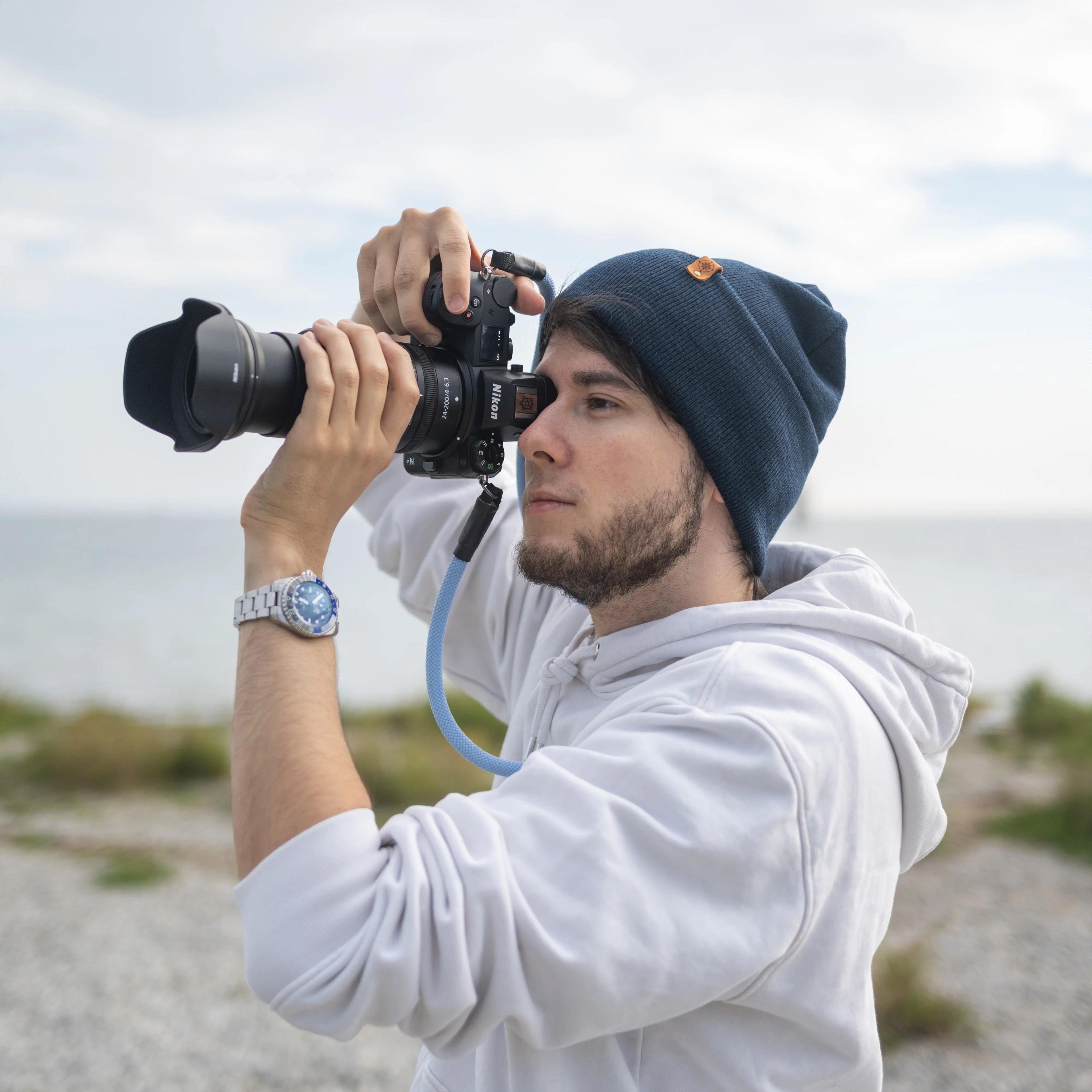 Junger Mann mit dunkelblauer Beanie-Mütze und weißem Hoodie fotografiert mit einer Spiegelreflexkamera am Meer. Die Mütze mit Kompass-Patch aus Kunstleder wurde nachhaltig veredelt in Deutschland – erhältlich bei nordkit.de.